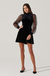 A woman is wearing a black velvet dress with sheer, puffed long sleeves that are gathered at the wrists with black cuffs. The dress has a high neckline and a fit-and-flare silhouette, ending above the knees. She is also wearing black high-heeled shoes with bows on the front.